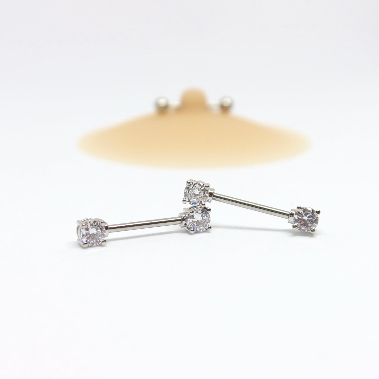 Pair of silver nipple piercing with clear gemstones placed in front of a pierced nipple cover by nip stories